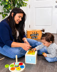 A woman and a toddler sit on a gray wood floor playing with a colorful wooden toy set from 123 Baby Box. The toy includes a small light blue and yellow drawer unit, peg rings, and a wooden mallet.