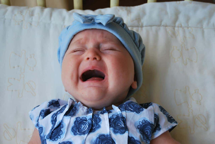 baby in a blue flower dress crying against a white pillow