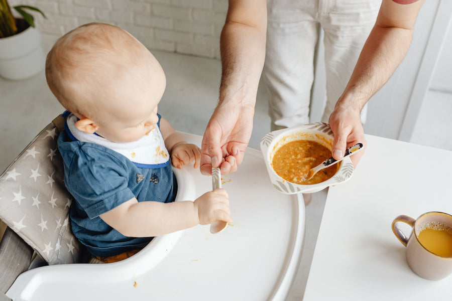 A person hands a spoon to a baby that sits in a high chair, while holding a bowl of soup in the other