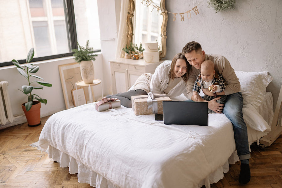 couple with a baby sitting on a bed with a present while looking at a laptop