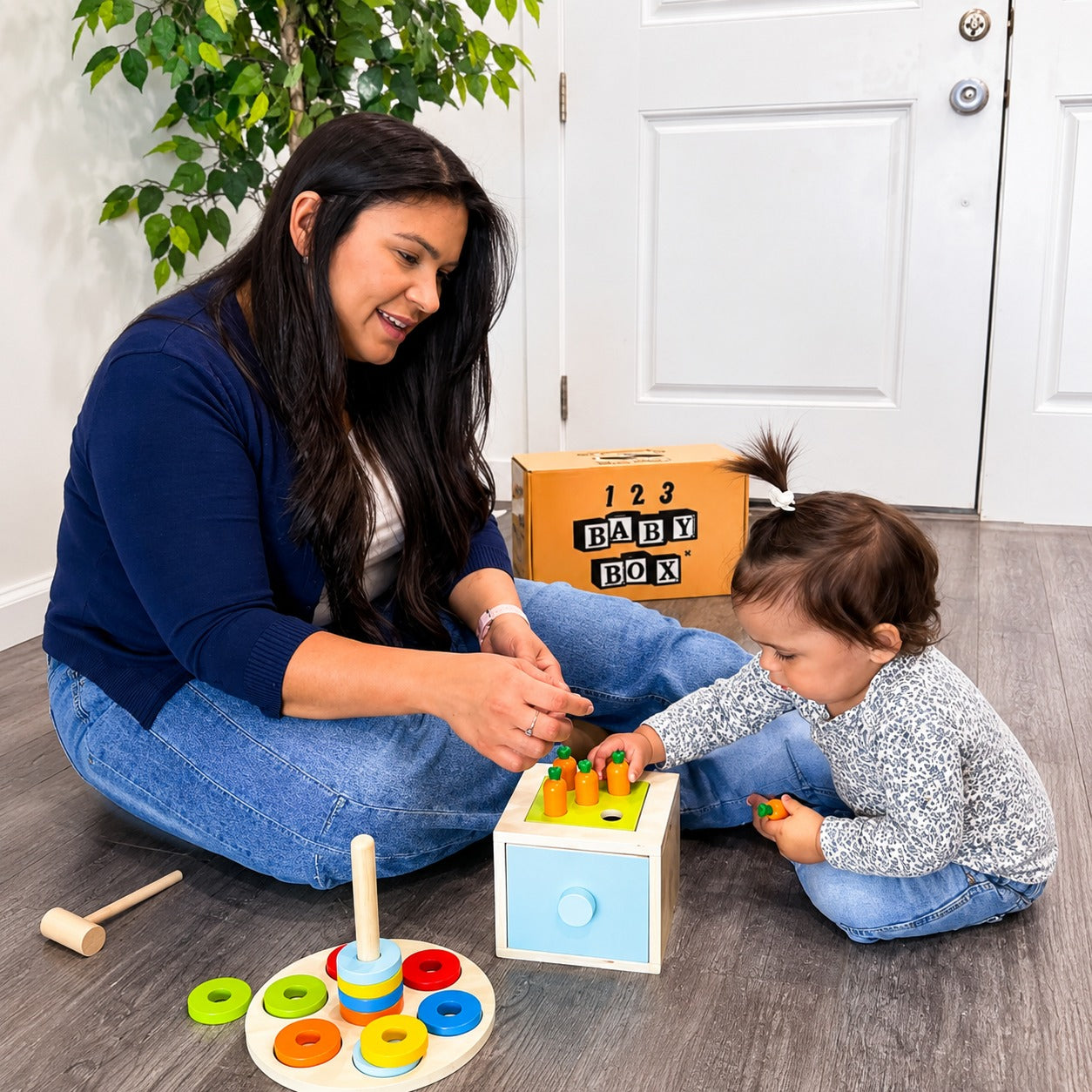 A woman and a toddler sit on a gray wood floor playing with a colorful wooden toy set from 123 Baby Box. The toy includes a small light blue and yellow drawer unit, peg rings, and a wooden mallet.